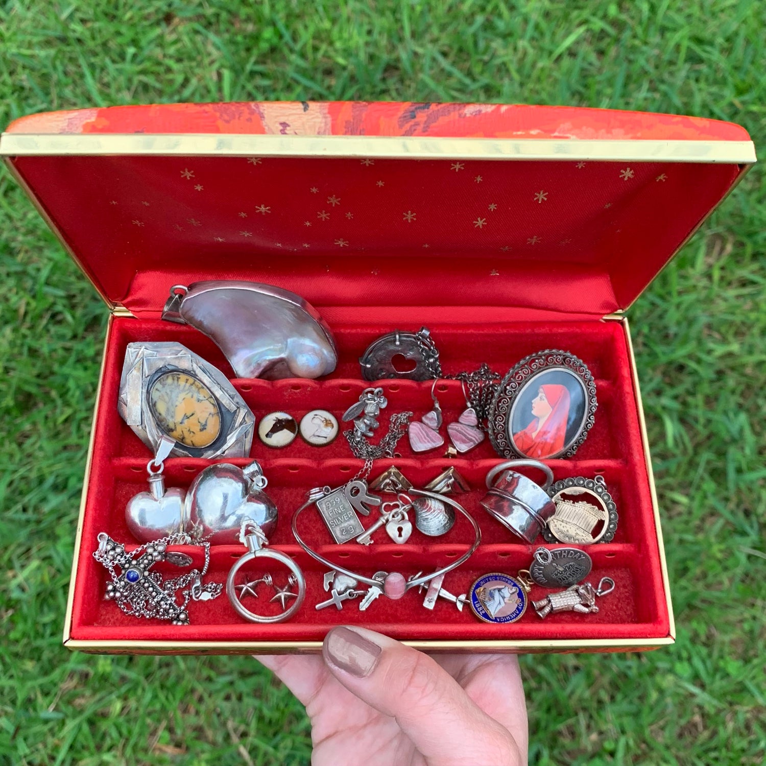 a hand holding open a red jewelry box showing an assortment of vintage jewelry